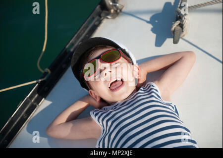 funny laughing small kid in captain hat lying on board luxury yacht laughing Stock Photo