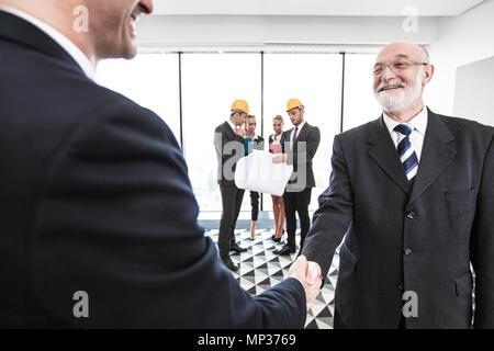 Business people at meeting shaking hands, their team in hardhats and blueprint on background, architects and investors Stock Photo