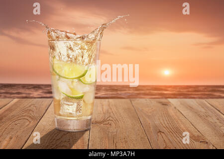 glass of cold splashing lemonade with lemon slices, mint leaves and ice cubes on wooden table against sea. Concept of rest and summer vacation Stock Photo