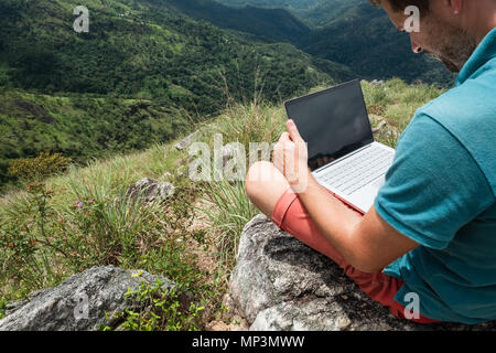 caucasian man with laptop sitting on the edge of ella mountain with stunning views of the valley in Sri Lanka. Stock Photo