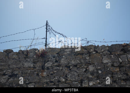 Stone wall with barbed wire against blue sky, wire torn and damaged Stock Photo