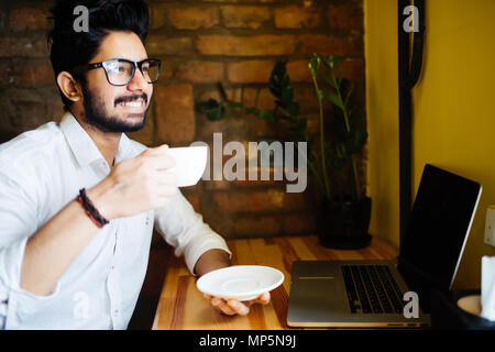Young Asian Indian businessman using computer during office break at cafe, relaxing with a cup of coffee. Stock Photo
