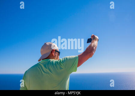 Back view of man web author is taking photo with cellphone camera for his web page in internet during trip, while is standing on a mountain against sky view background with copy space Stock Photo