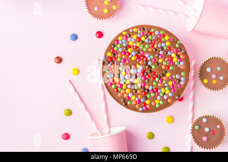 Milk Chocolate Birthday Cake with Multicolored Glazed Candy Sprinkles Buttercups Paper Drinking Cups Pink Straws Pink Background. Party Celebration Ki Stock Photo