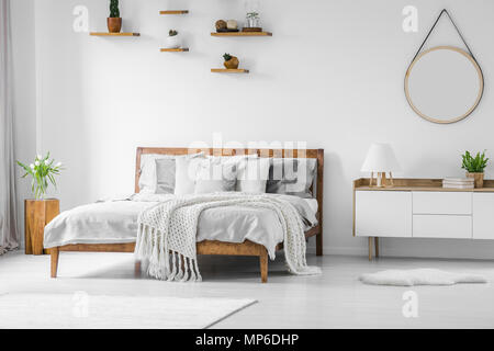 Comfortable, big, wooden bed with linen, pillows and blanket, nightstand beside and round mirror hanging on a white wall in a bright bedroom interior. Stock Photo