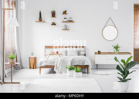 Morning in a bright and sunny modern white bedroom interior with wooden furniture. Cushions, blanket and food tray on the bed, nightstand beside and h Stock Photo