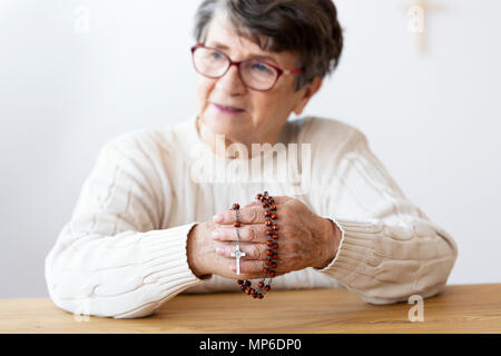 Religious senior woman holding red rosary with cross while praying. Focus on the hands Stock Photo