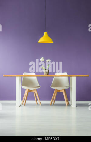 Symmetric, modern, minimal style interior with a front view of two white chairs behind a table with a flower in a pot and a yellow lamp against a purp Stock Photo