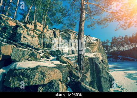 Rocky shore of the lake in winter. The frozen lake in the forest. Pines grow on a rock Stock Photo