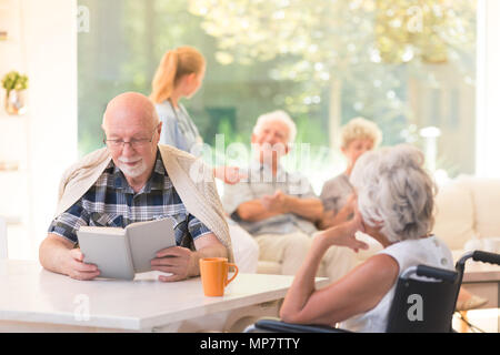 Senior man reading a book and disabled woman sitting i a wheelchair at a table and drinking tea Stock Photo