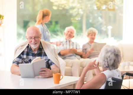 Senior man reading a book and smiling while sitting with a friend at a table in nursing house Stock Photo