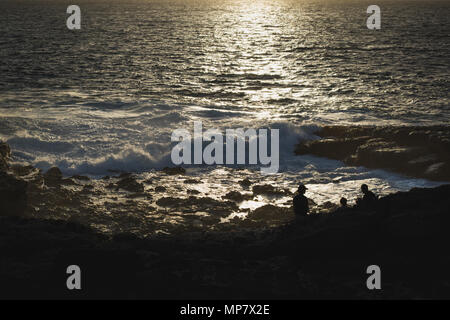 Sunset on Atlantic Ocean with three people enjoying it sitting on rocks. Stock Photo