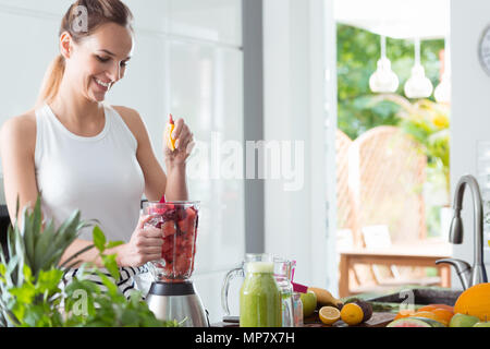 Smiling woman squeezing orange juice into blender with sliced watermelon and beet while preparing smoothie in kitchen Stock Photo