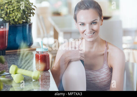 Smiling fit woman sitting on chair next to kitchen countertop with fruit cocktail and green dumbbells Stock Photo