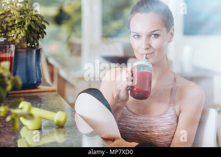 Satisfied fit woman drinking red smoothie after fitness class in kitchen with green dumbbells on countertop Stock Photo