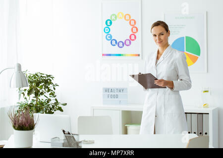 Professional nutritionist in white uniform in the office with plant and colorful posters Stock Photo