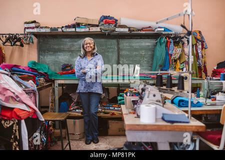 Smiling mature seamstress standing in her busy sewing workshop Stock Photo