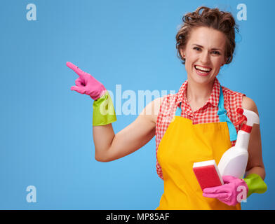 Big cleaning time. happy modern woman in a yellow apron with kitchen sponge and a bottle of detergent pointing at something on blue background