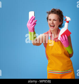 Big cleaning time. happy young woman with rubber gloves and kitchen sponge and a bottle of detergent taking selfie with mobile phone against blue back Stock Photo