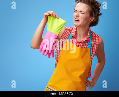 Big cleaning time. sad young housewife in a yellow apron sniffing rubber gloves isolated on blue Stock Photo