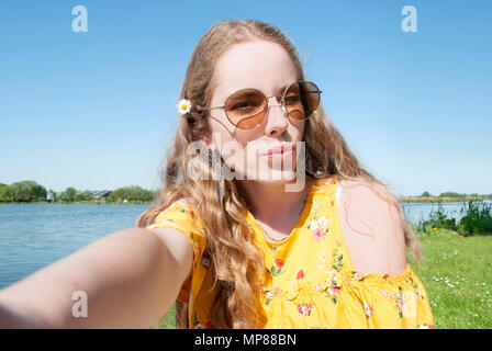 beautiful young millennial girl, taking selfie pcture with cell phone camera, outdoors in park on sunny day Stock Photo