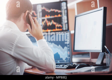 Over the shoulder view of and stock broker trading online while accepting orders by phone. Multiple computer screens ful of charts and data analyses in background Stock Photo
