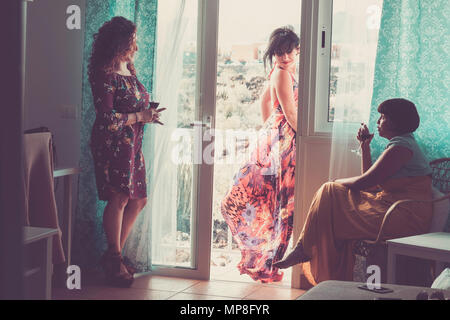 three caucasian middle age friends at home in an usual day. two inside and one outsdide in the terrace speaking together drinking a cup of wine Stock Photo