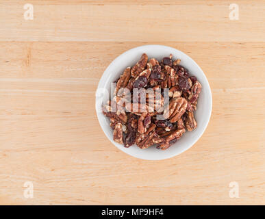 Top view of a small white bowl filled with sugar glazed pecans and dried cranberries on a wood table. Stock Photo