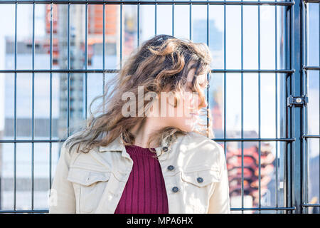 Side profile of peaceful young woman by urban fence, in sun sunlight, warm in New York City Hudson Yards Stock Photo