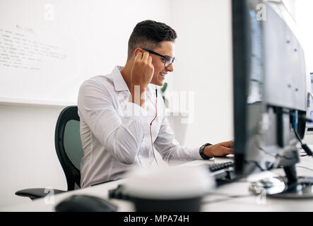 Smiling software developer sitting at his office desk working on laptop wearing earphones. Man wearing spectacles working on laptop computer in office Stock Photo