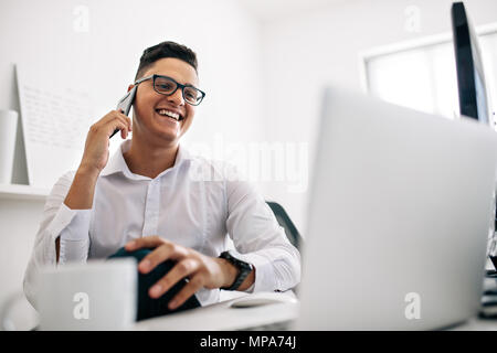 Smiling software developer talking over mobile phone sitting at his office desk with a laptop and coffee cup in front. Man wearing spectacles working  Stock Photo