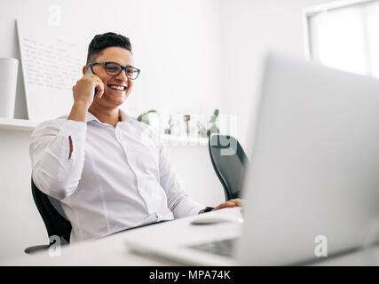 Smiling software developer talking over mobile phone sitting at his office desk with a laptop in front. Man wearing spectacles working on laptop compu Stock Photo