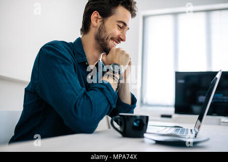 Software developer sitting in front of computer and working in office. Smiling man looking at laptop sitting with his chin resting on hands with a cof Stock Photo