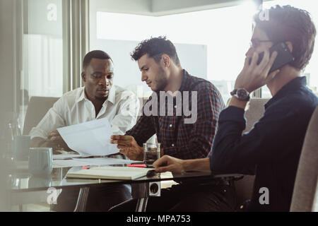 Businessmen discussing work sitting at conference table in office. Two men discussing work while another man is talking on mobile phone. Stock Photo