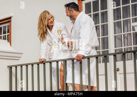 Couple wearing white bath robes standing on balcony and smiling at each other. Smiling man and woman in bathrobes having wine and talking. Stock Photo