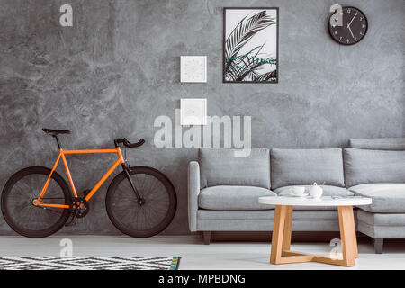 Orange bike next to grey sofa in living room with coffee table,black clock and posters on concrete wall Stock Photo