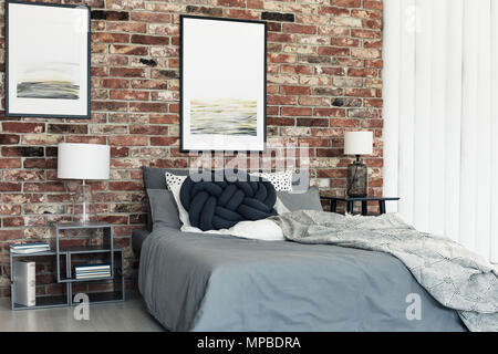 Black knot pillow on bed with grey bedding in bedroom with lamps and posters on brick wall Stock Photo