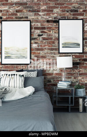 Paintings on brick wall above bed and cabinet with lamp in modern bedroom interior Stock Photo