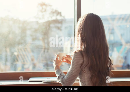 the woman in the cafe sits at a table, drinks coffee and looks at the panorama of the city outside the window. A girl in a cafe on a sunny morning Stock Photo