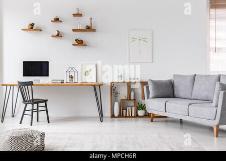 Grey sofa near black chair at desk with computer monitor in open space interior with poster. Real photo Stock Photo
