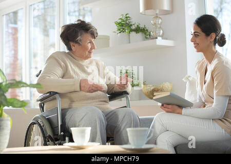 Professional caregiver in uniform reading a book and talking to a happy senior woman in a wheelchair during leisure time in luxury nursing home common Stock Photo
