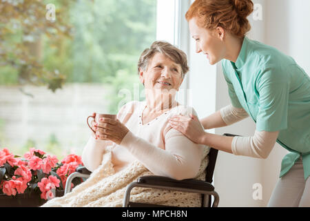 A professional caretaker in uniform helping a geriatric female patient on a wheelchair. Senior holding a cup and sitting by a large window in a rehabi
