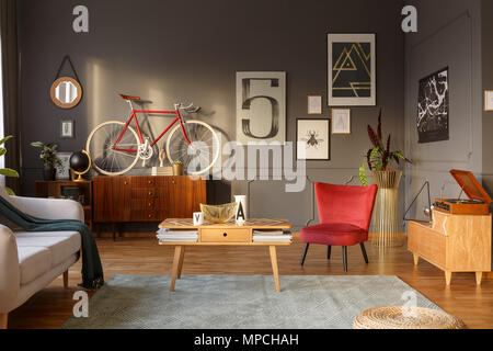 Sun shining into a unique, comfortable living room interior with couch, table, red armchair, green carpet, bicycle, gramophone and intriguing posters  Stock Photo