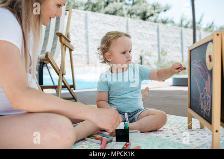 A little boy and his mother sitting on a patio and drawing on a chalkboard. A mother admiring first drawings of her son on a chalkboard. Stock Photo