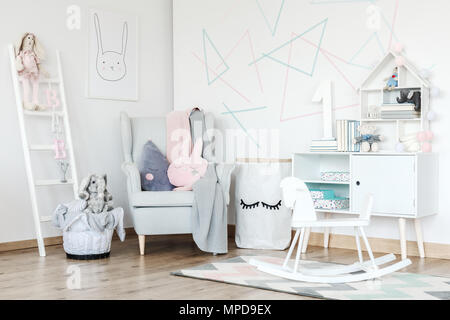 Pink pillow on grey armchair and white rocking horse on carpet in scandi kid's room with cupboard Stock Photo