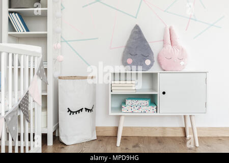 Pink and grey pillow on white cupboard in kid's room with paper bag and shelf against triangles wall Stock Photo
