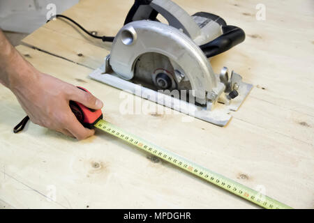 Man's hand in working glove measures measuring tape on  background of plywood circular saw blade Stock Photo