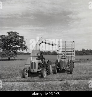 1950s, historical, male farmer on tractor with attached combine or harvester, with another farmer and tractor with a wooden-slatted trailer attached, beside him, to catch the harvested crop, England, UK. In this era, with post-war restrictions still in place, farming was an important industry and - difficult to believe today - most of the land mass of Britain was still predominantly rural and agricultural. Stock Photo