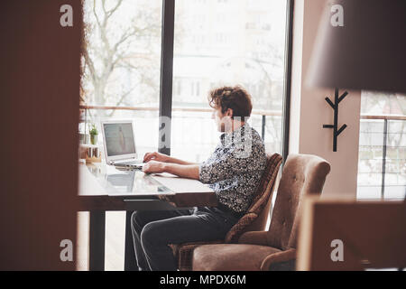 Young handsome man sitting in office with cup of coffee and working on project connected with modern cyber technologies. Businessman with notebook trying to keep deadline in digital marketing sphere Stock Photo