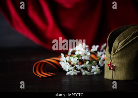 Concept, greeting card to the Victory Day. Soldier's cap with red star, cherry blossom and guards ribbon on the background of the red standard Stock Photo